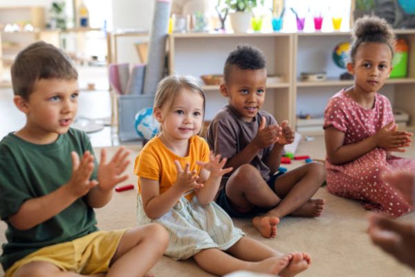 A group of small nursery school children sitting on floor indoors in classroom, clapping.